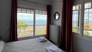 A bedroom with a large bed, two hanging pendant lights, a view of the sea, and a window with curtains. - Villa Eleni Panorama Saronida (Saronida)