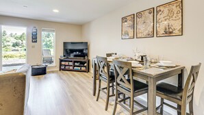 A dining area with a wooden table, chairs, and a television. There are framed pictures on the wall and a bookshelf with books and decorative items. - Morning Hikes & Beach Nights: Canalfront Condo! (Ocean Shores)