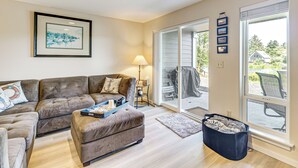 A living room with a brown sofa, a matching ottoman, a framed picture on the wall, and a large sliding glass door leading to an outdoor area. - Morning Hikes & Beach Nights: Canalfront Condo! (Ocean Shores)