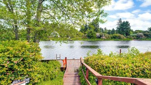 A wooden deck leading to a calm lake surrounded by lush greenery and trees. - Morning Hikes & Beach Nights: Canalfront Condo! (Ocean Shores)