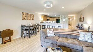 A modern living room with a sofa, dining table, and chairs. A kitchen is visible in the background with a refrigerator and microwave. - Morning Hikes & Beach Nights: Canalfront Condo! (Ocean Shores)