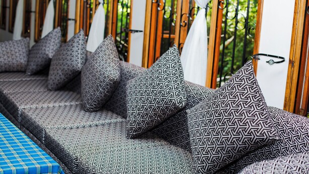 A row of cushioned seating with patterned pillows, a blue checkered table, and a view of greenery through a window with a wooden frame. - Kili Share Hotel & Hotel (Himo)