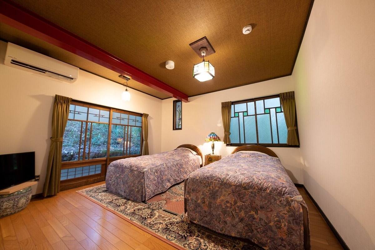 Ein Zimmer mit zwei Betten, einem Fernseher, einem Fenster mit Vorhängen und einem Deckenventilator.
