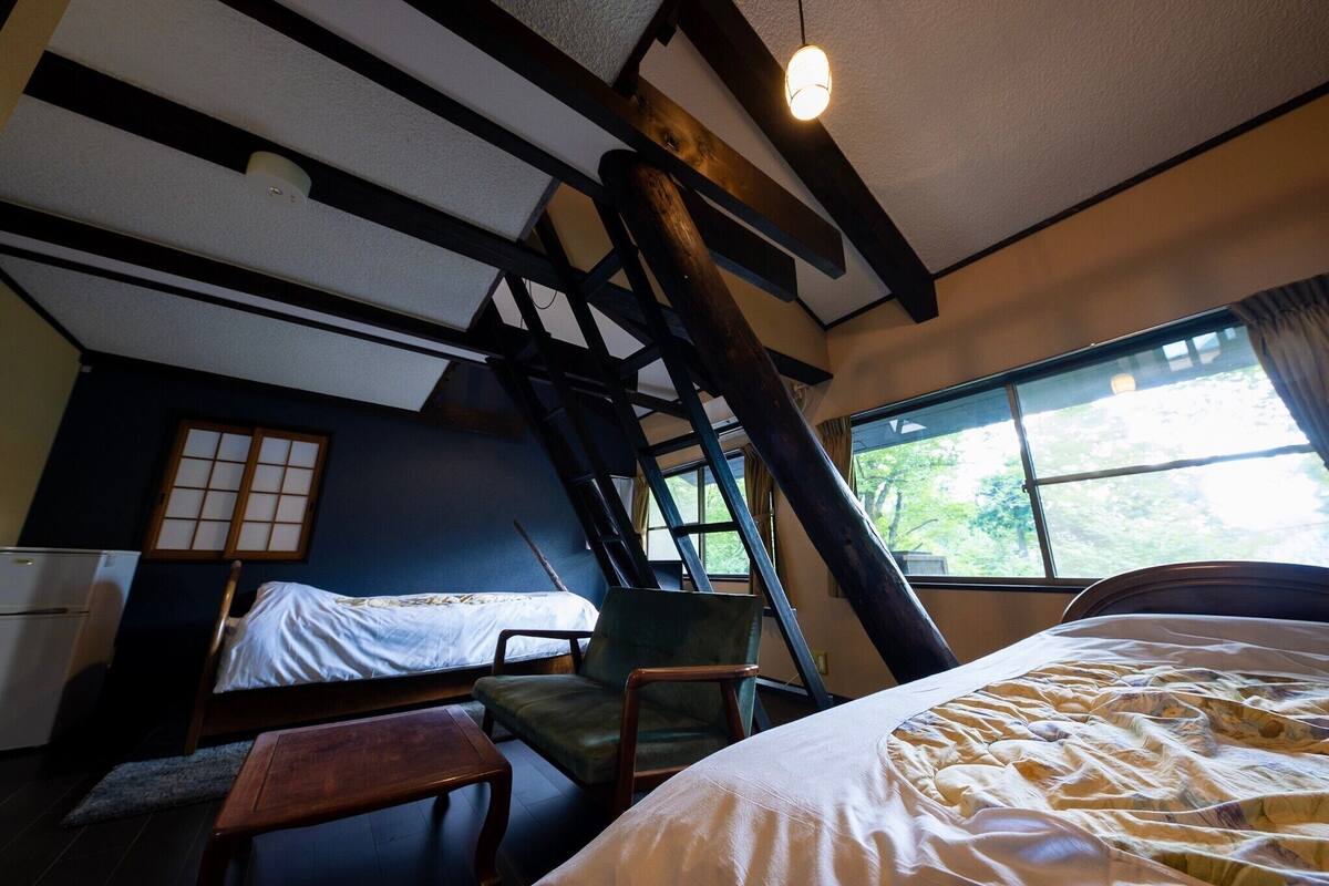Ein Zimmer mit schräg laufender Decke, einem Bett, einem Stuhl, einem kleinen Tisch und einem Fenster mit Blick auf Grünflächen.