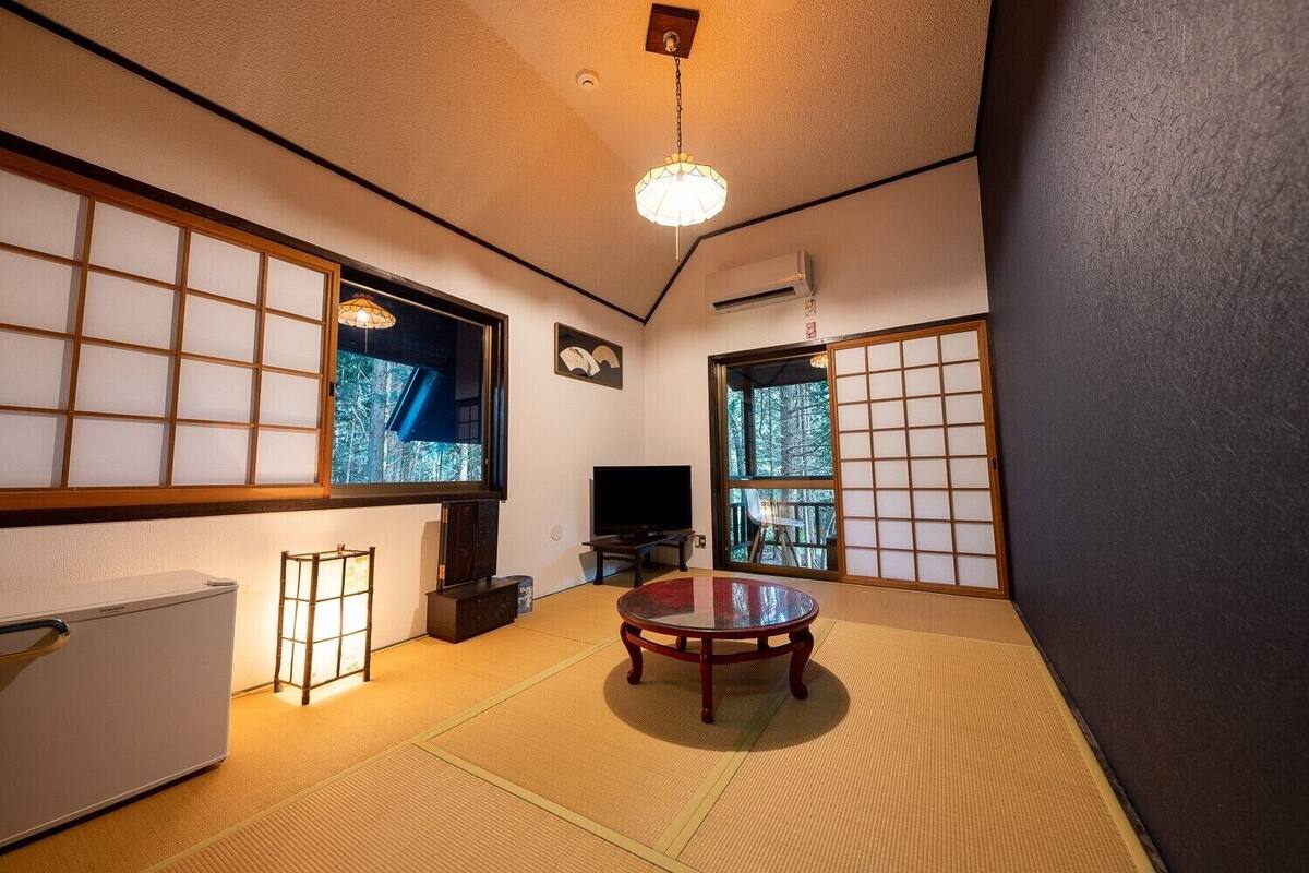 Ein traditioneller japanischer Raum mit Tatami-Bodenbelag, einem niedrigen Tisch, einem Fernseher und einer Schiebetür mit Blick auf Bäume.