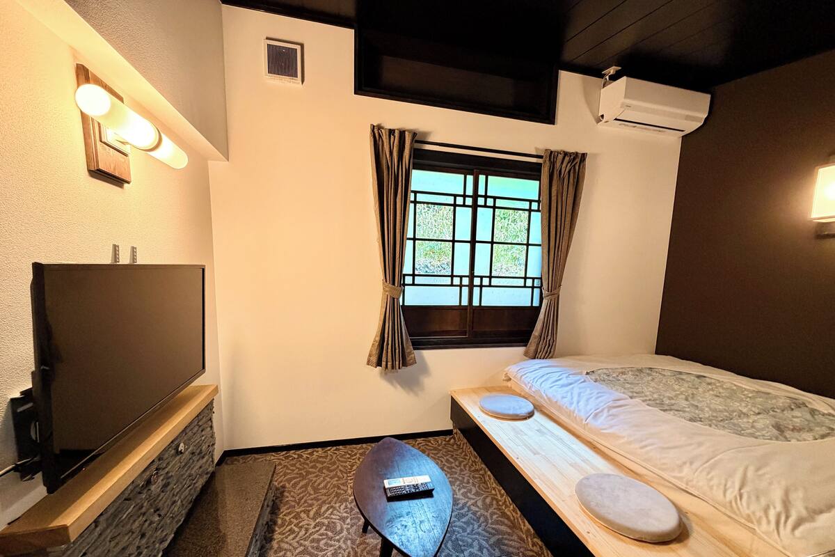 Ein Schlafzimmer mit einem Bett, einem Fernseher, einem kleinen Tisch und einem Fenster mit Vorhängen.
