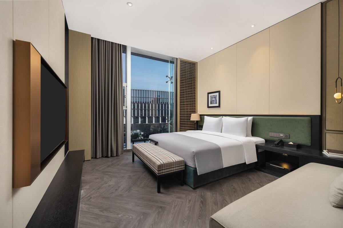 Une chambre d’hôtel moderne avec un grand lit, une banquette, une table de chevet et une vue sur la ville, visible à travers une porte vitrée.