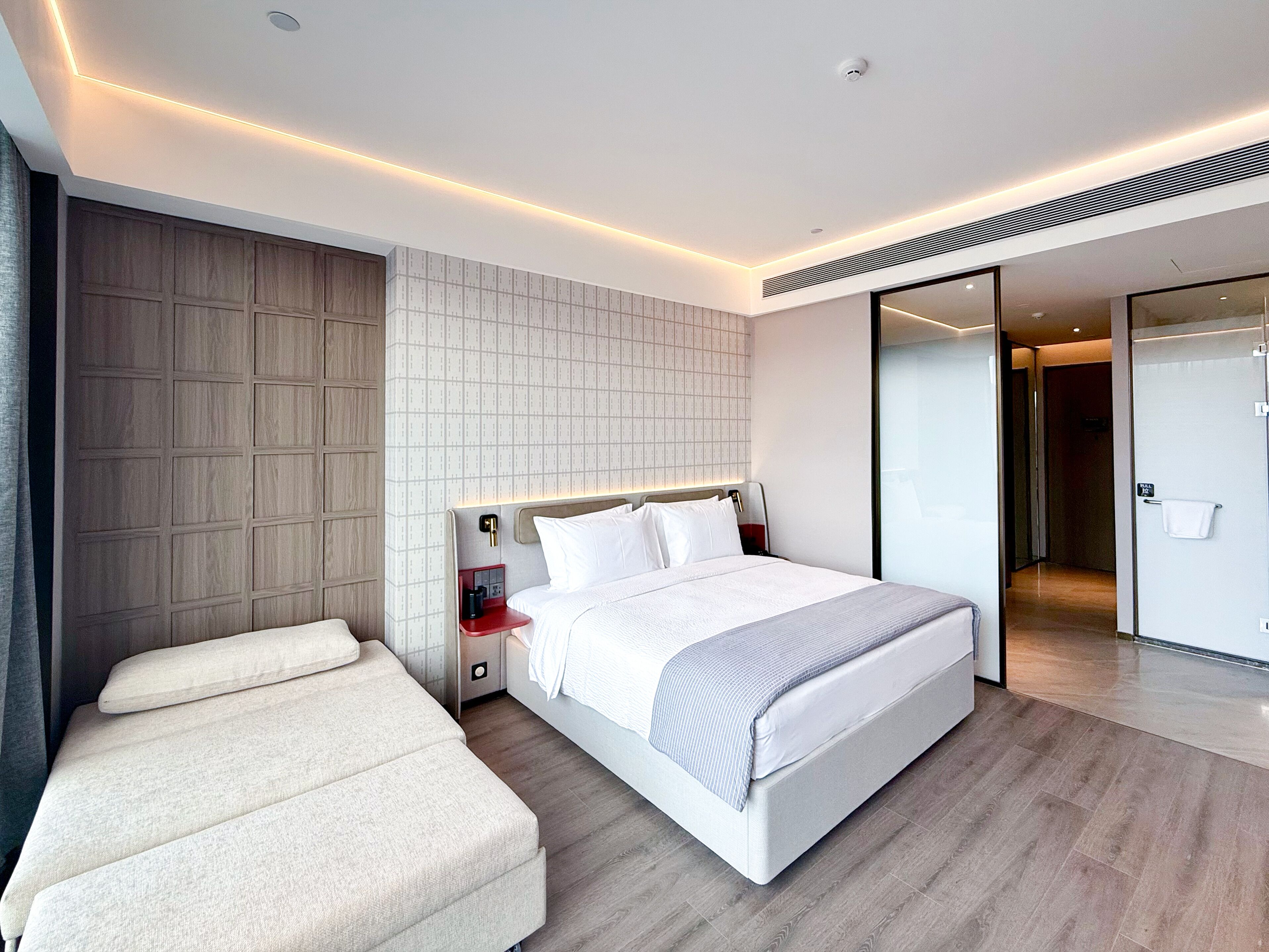 Een moderne hotelkamer met een groot bed, een nachtkastje, een inbouwkbank en een schuifdeur naar een badkamer.