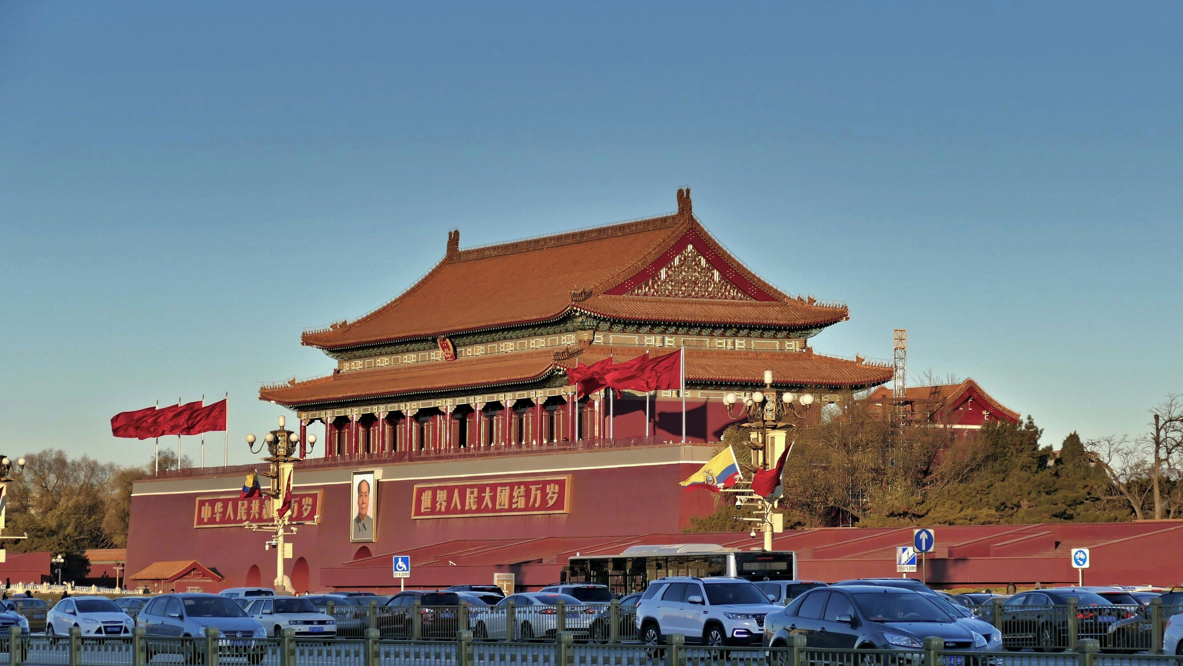 A large traditional building with a red roof and multiple flags, surrounded by a parking area with cars.
