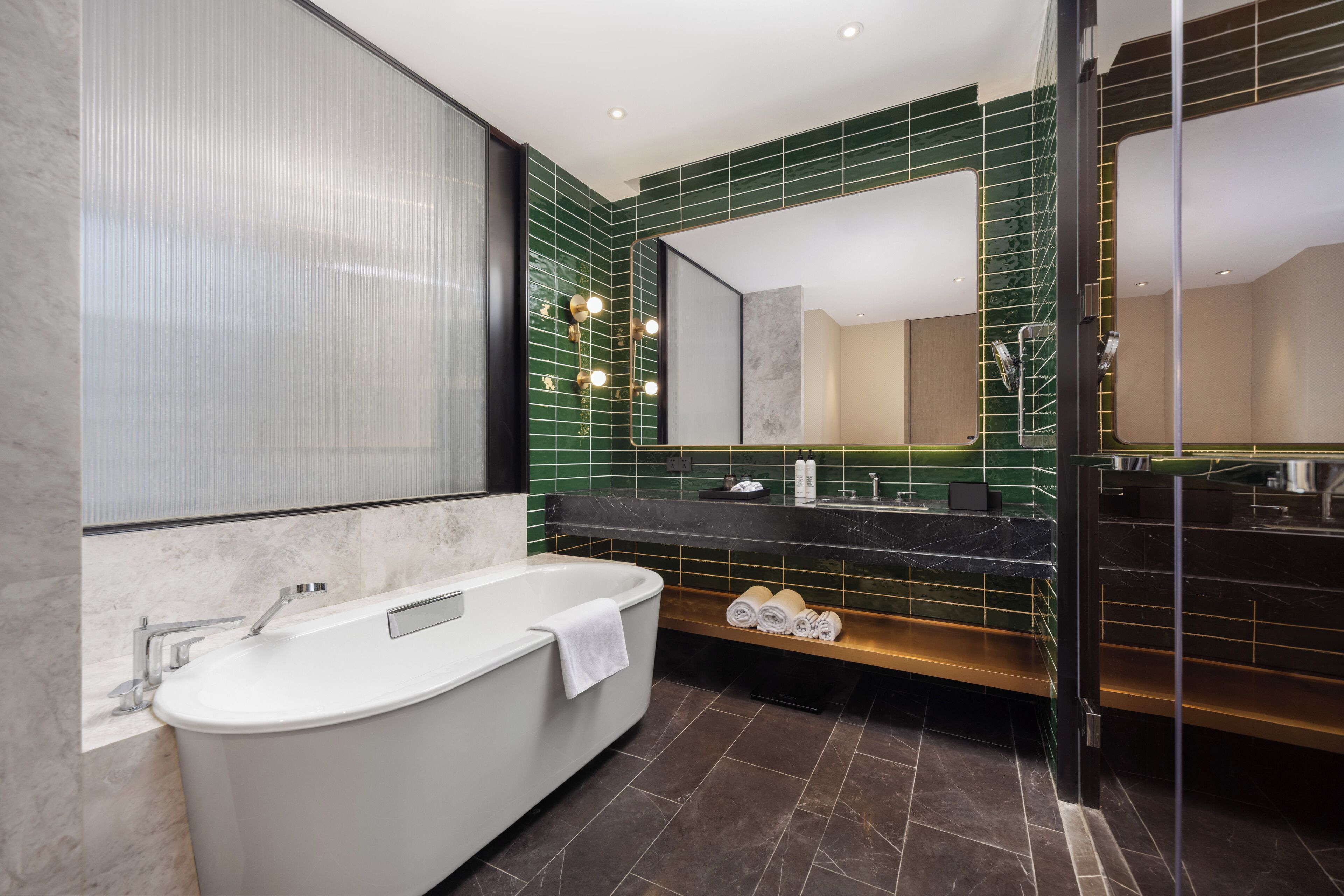 A modern bathroom with a freestanding bathtub, a large mirror, and a green-tiled wall.
