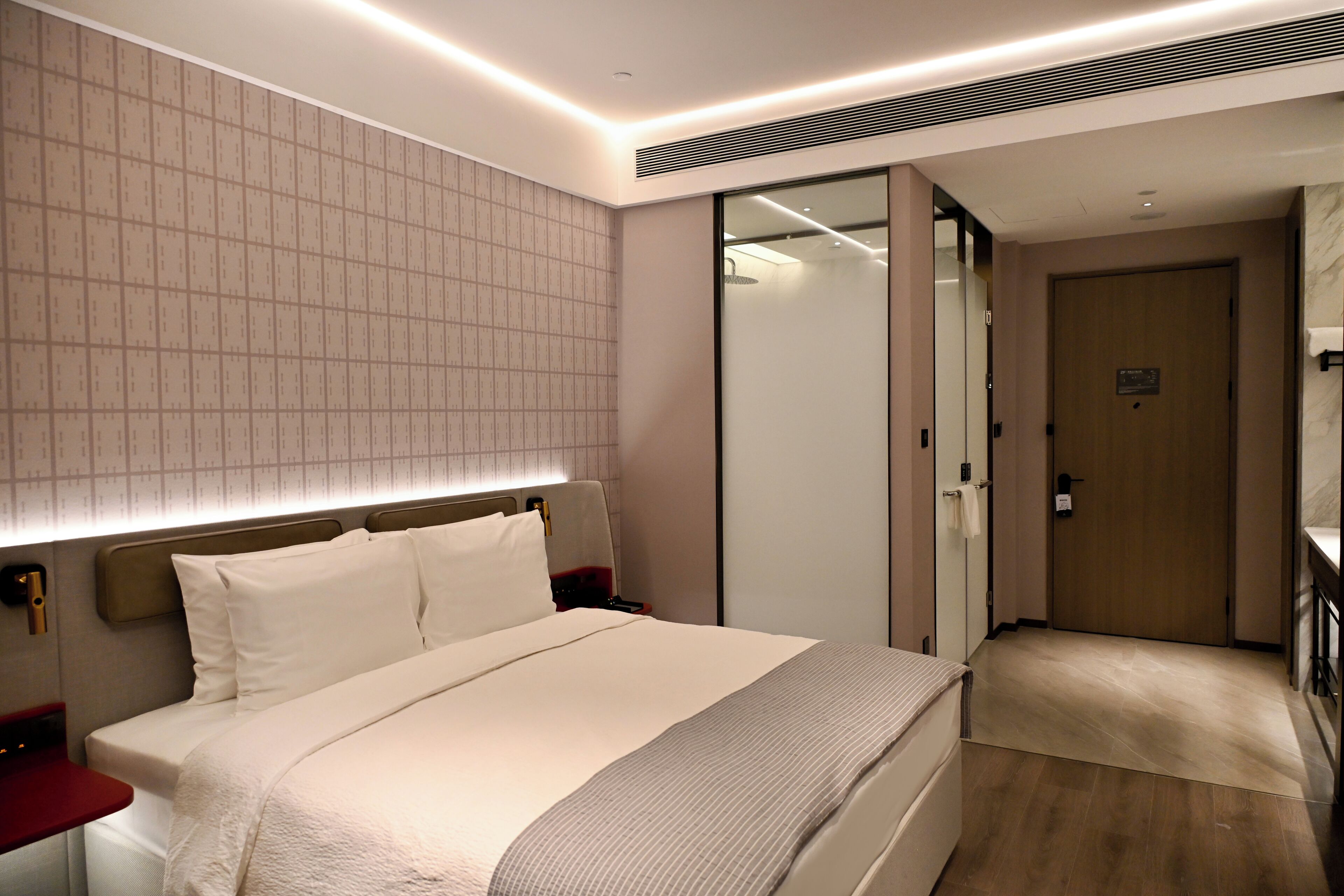 Una habitación de hotel moderna con una cama grande, mesitas de noche, un armario y un baño con puerta de cristal.