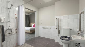 Une salle de bain moderne avec douche, toilettes et lavabo, un lit dans la pièce adjacente et une barre d’appui visible.