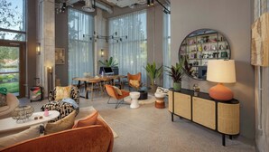 Una habitación de hotel moderna con un ventanal grande, un comedor, una zona de estar con sofá y sillones, una mesita y un espejo decorativo.