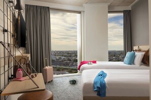 A hotel room with two beds, a desk with a lamp, a suitcase, and a view of a cityscape. - Moxy Dublin Docklands (Dublin)