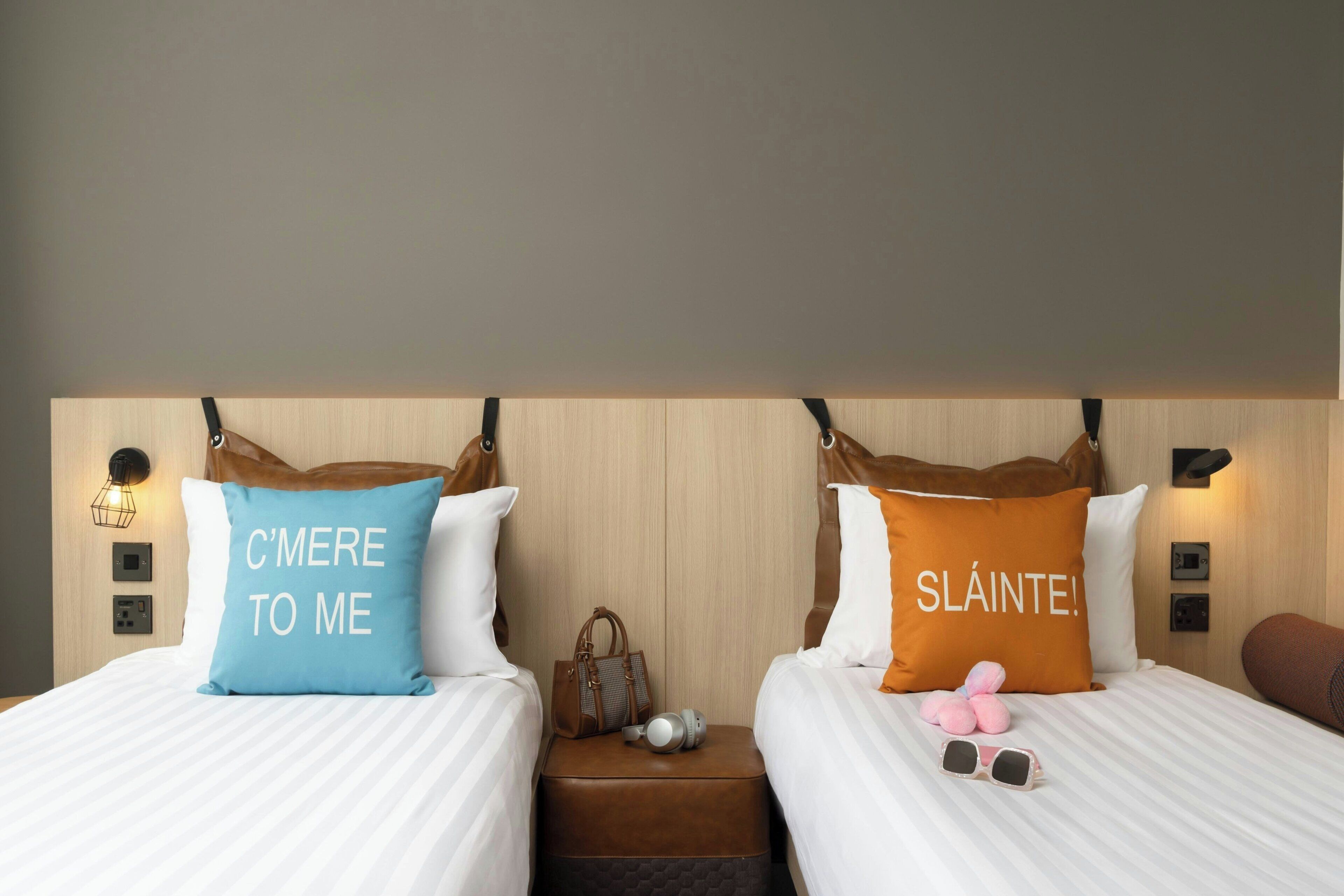 Une chambre d’hôtel avec deux lits, chacun étant agrémenté d’un oreiller orné d’une phrase en français.