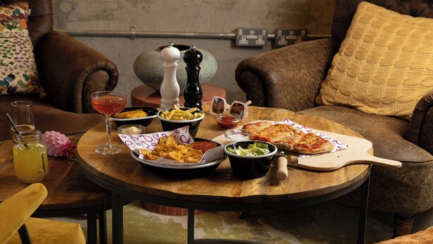 A round wooden table with a variety of food items, including pizza, fries, and salad, set on a couch with brown leather cushions.