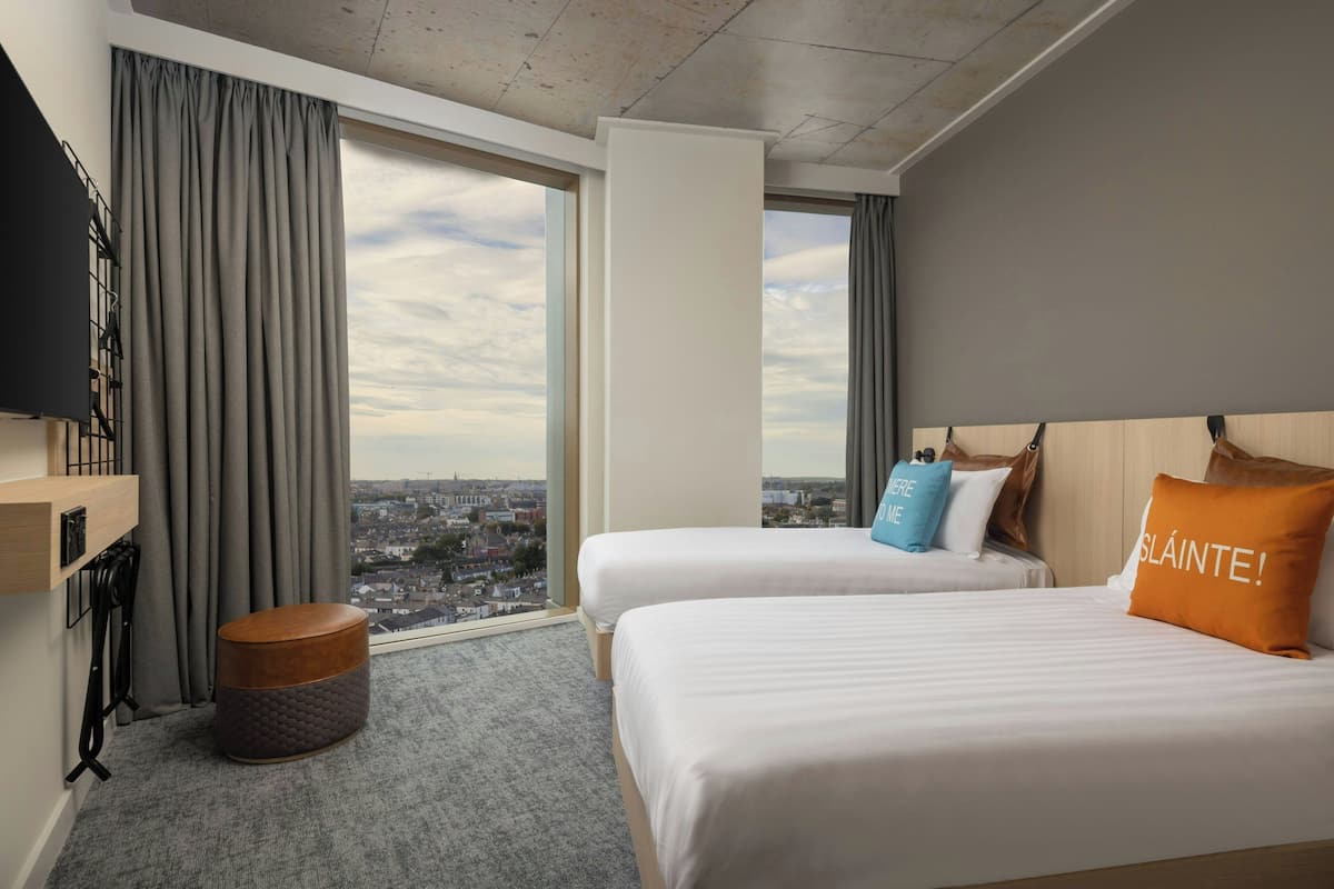 Ein Hotelzimmer mit zwei Betten, einem großen Fenster mit Blick auf die Stadt, einem kleinen Hocker und einem an der Wand befestigten Fernseher.