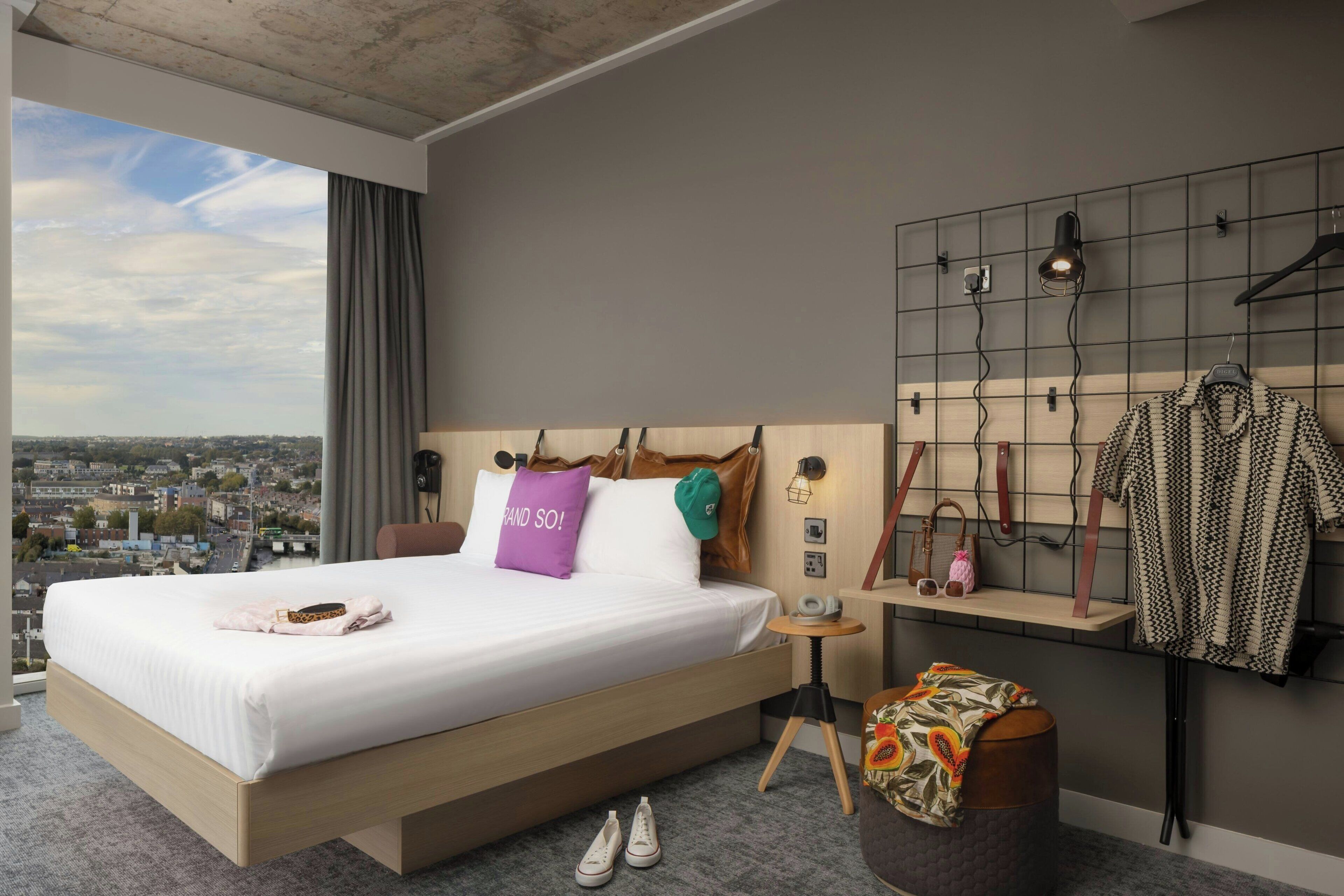 A hotel room with a large bed, a view of the city, a wall-mounted light, and a small table with a phone and a pair of shoes.