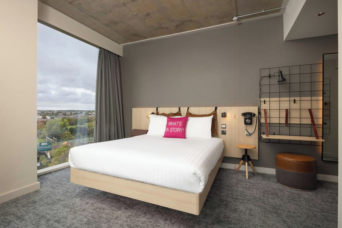 Ein modernes Hotelzimmer mit einem großen Bett, Blick auf die Stadt, einem kleinen Tisch und einem an der Wand befestigten Regal.