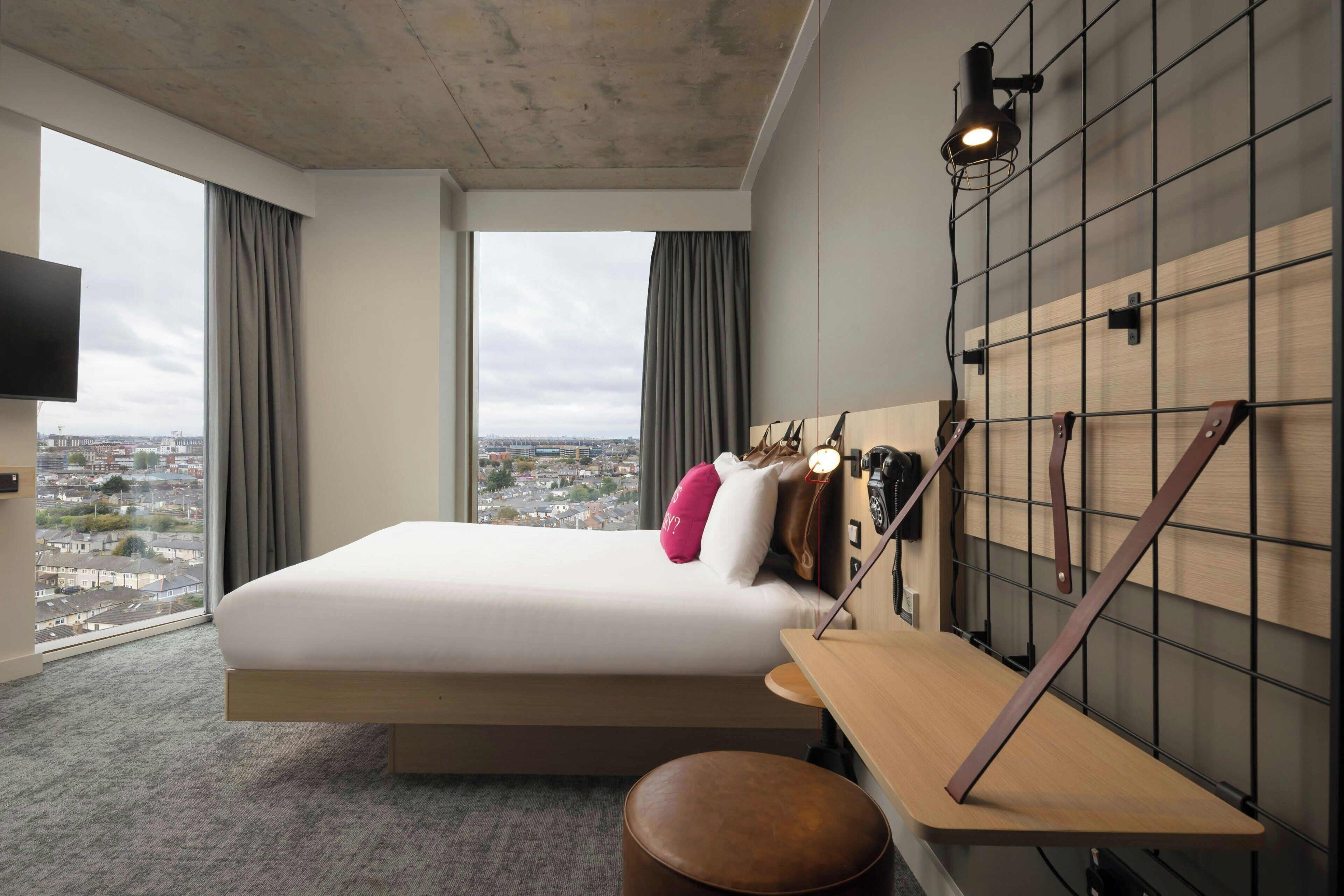 Quarto de hotel moderno com cama grande, escrivaninha, cadeira, televisão e vista para a cidade.