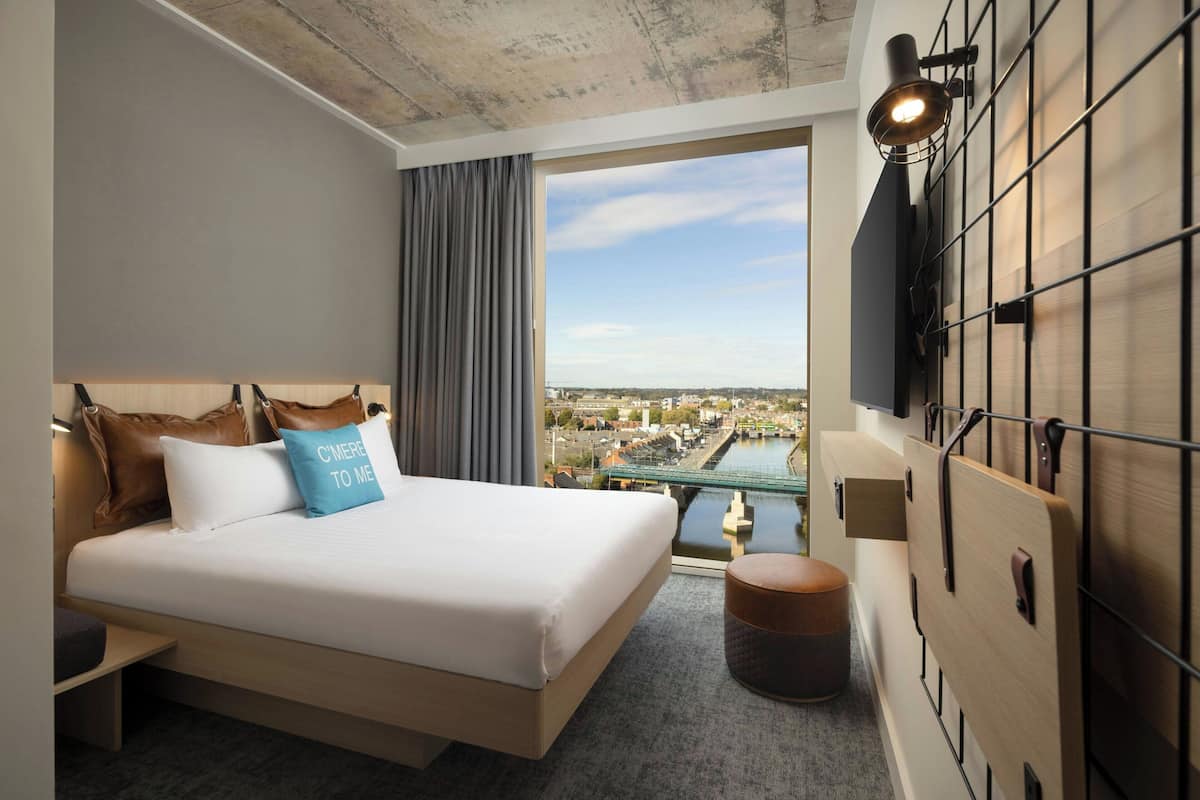 Ein Hotelzimmer mit Bett, Blick auf die Stadtlandschaft, einem an der Wand befestigten Fernseher und einem kleinen Hocker.