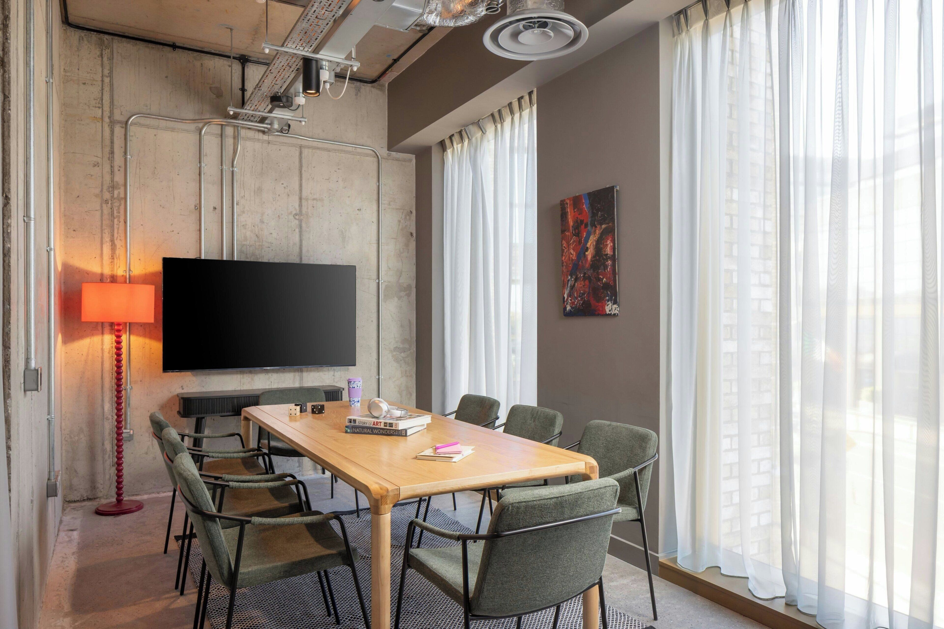 Ett konferensrum med ett långt bord, sex stolar, en platt-TV, en röd golvlampa och ett stort fönster med tunna, genomskinliga gardiner.
