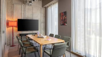 Un salón de conferencias con una mesa larga, seis sillas, un televisor de pantalla plana, una lámpara de pie roja y un ventanal con cortinas traslúcidas.