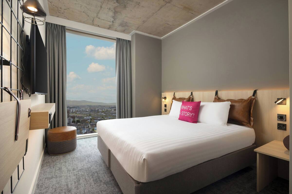 Ein modernes Hotelzimmer mit einem großen Bett und Blick auf die Stadt, wobei sich ein Stadtbild durch das Fenster zeigt.