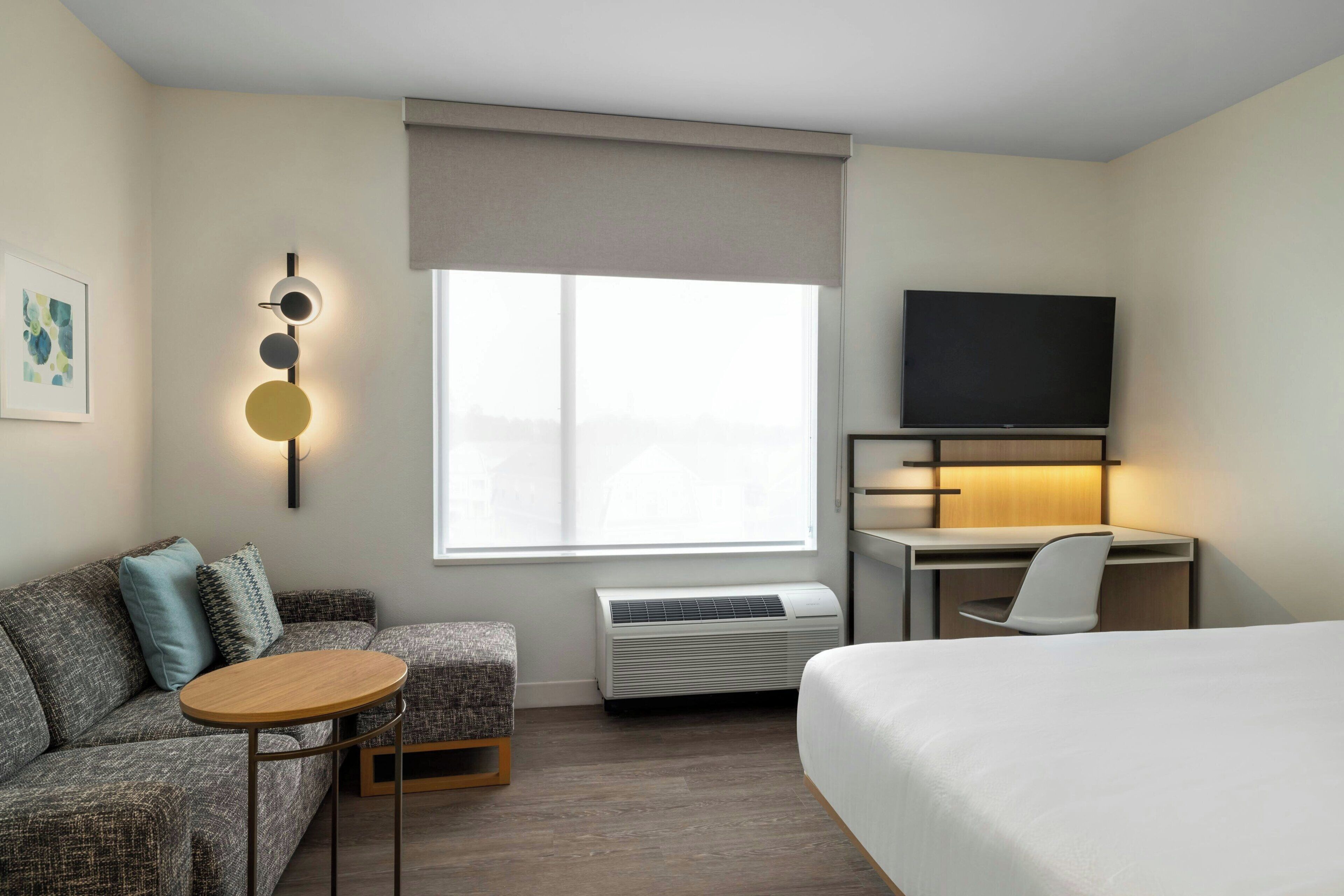 Habitación de hotel con cama, escritorio, silla, mesita, sofá, aire acondicionado, televisor y ventana con vistas.