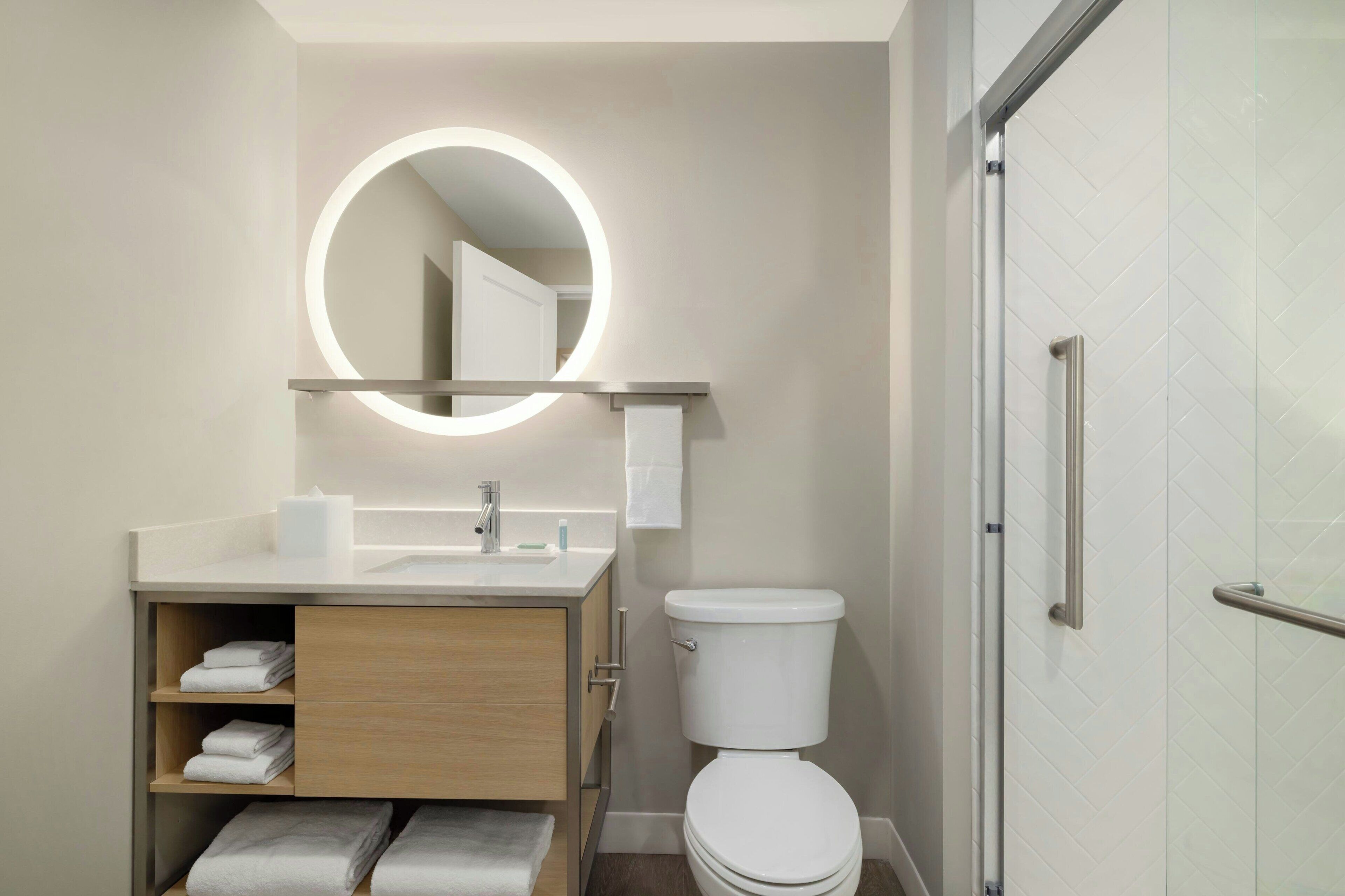 A modern bathroom with a toilet, sink, and mirror. There are towels on a shelf, and a shower area is visible.