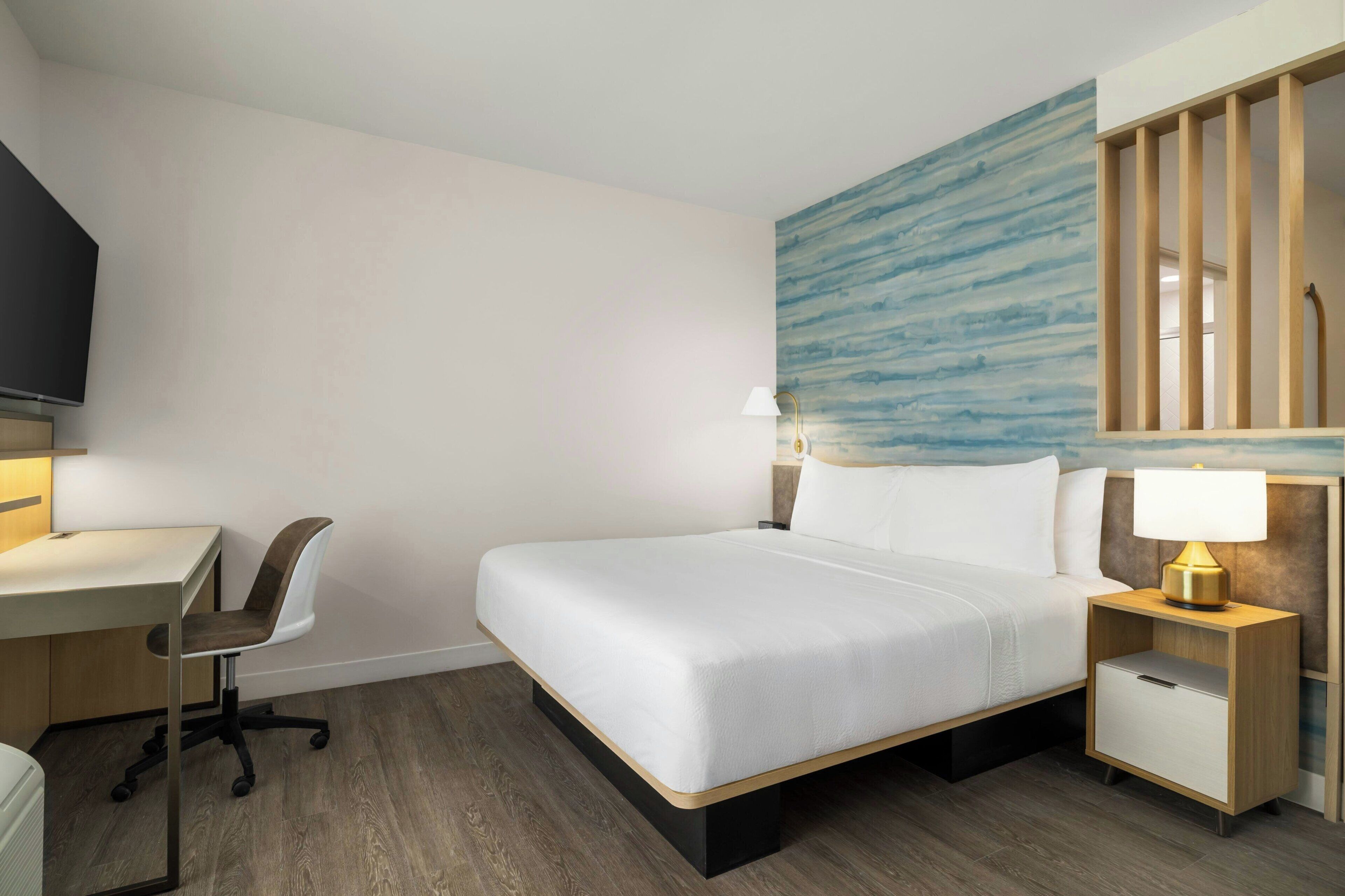 A hotel room with a bed, a desk, a chair, a nightstand, and a wall with a blue abstract pattern.