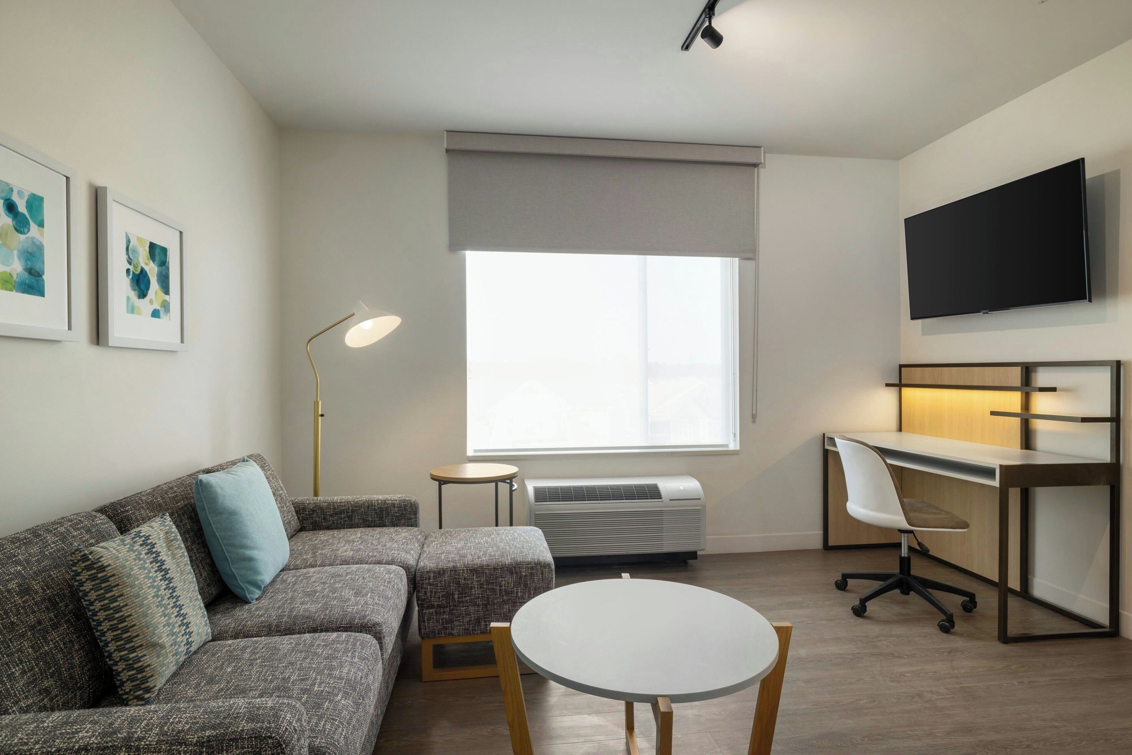 Ein modernes Wohnzimmer mit grauem Sofa, einem runden Tisch, einer Stehlampe, einem an der Wand befestigten Flachbildfernseher und einem Fenster mit grauen Jalousien.