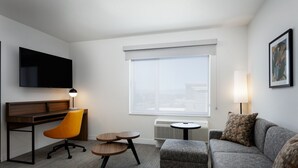 Een moderne hotelkamer met een flatscreen televisie, een bureau met stoel, een grijze bank, een klein rond tafeltje en een groot raam met uitzicht.