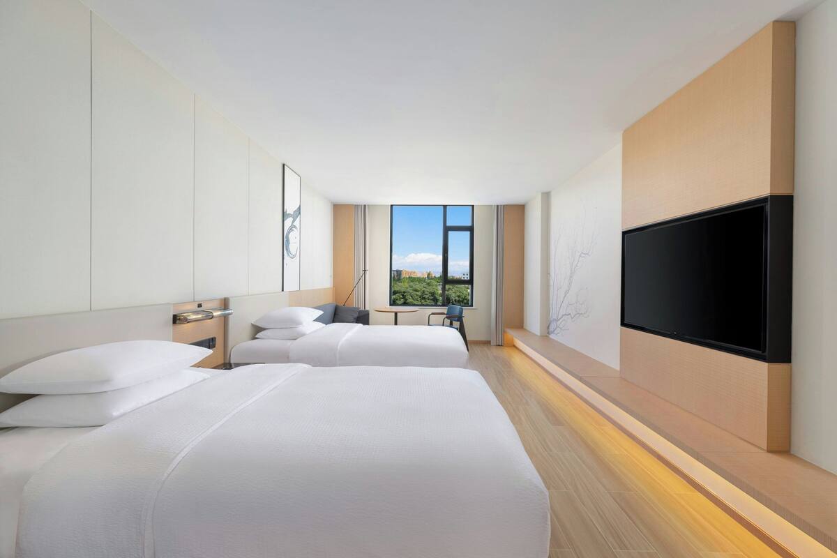 Ein modernes Hotelzimmer mit zwei Betten, einem Flachbildfernseher, einem Fenster mit Blick auf die Stadt und einem kleinen Tisch mit Stuhl.