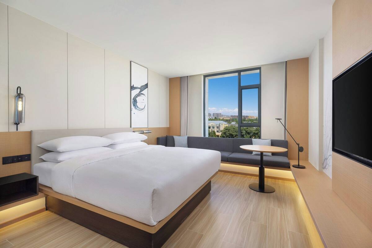 Ein modernes Hotelzimmer mit einem großen Bett, einer Sitzecke mit Tisch, einem Fernseher und Blick auf die Stadt.