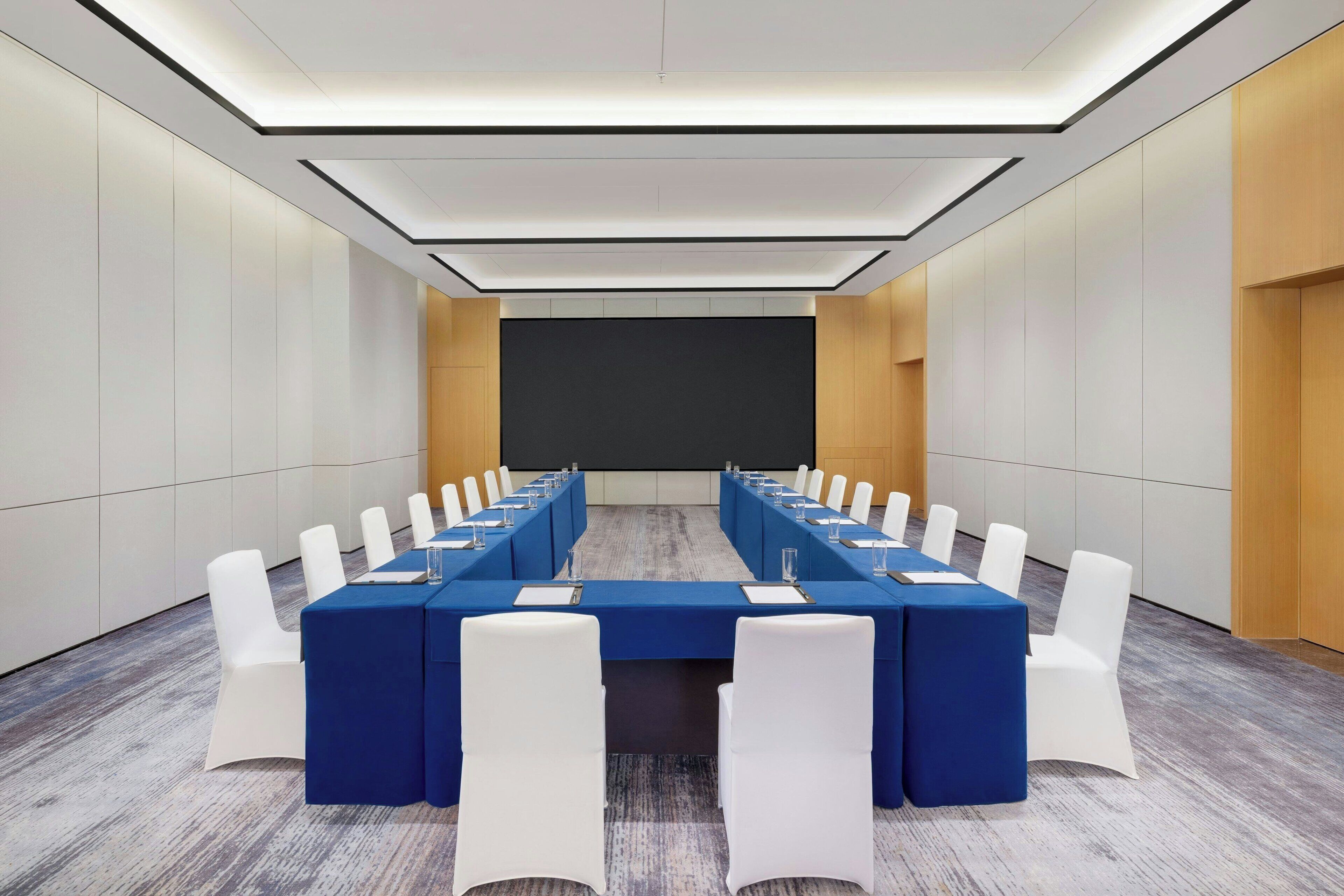 A conference room with a long table, white chairs, and a projector screen.