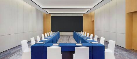 Ein Konferenzraum mit einem langen Tisch, weißen Stühlen und einer Projektorleinwand.