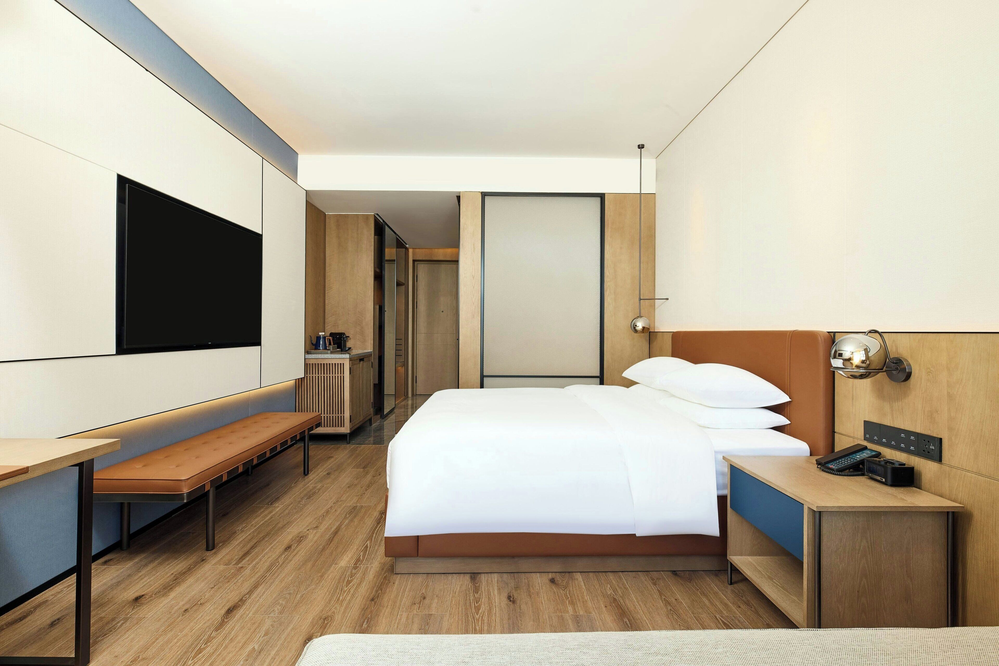 Una habitación de hotel moderna con cama, escritorio, banco, televisor y mesita de noche con lámpara.