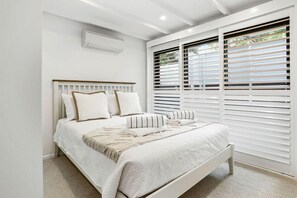 A bedroom with a white bed, white headboard, and white blinds on the windows. - Coastal Luxe Little Cove (Noosa Heads)