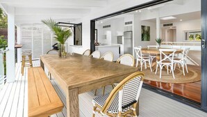 Un vaste espace de salle à manger avec une table en bois et des chaises en osier, attenant à une cuisine et à un balcon doté de balustrades blanches.