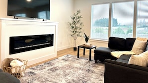 A modern living room with a flat-screen TV mounted above a fireplace, a large window with blinds, and a cozy seating area with a dark-colored sofa and cushions.