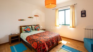 A bedroom with a bed, a nightstand, a window with curtains, a hanging light fixture, and a radiator. - Infinity Waves by ACasadasCasas (Ericeira)