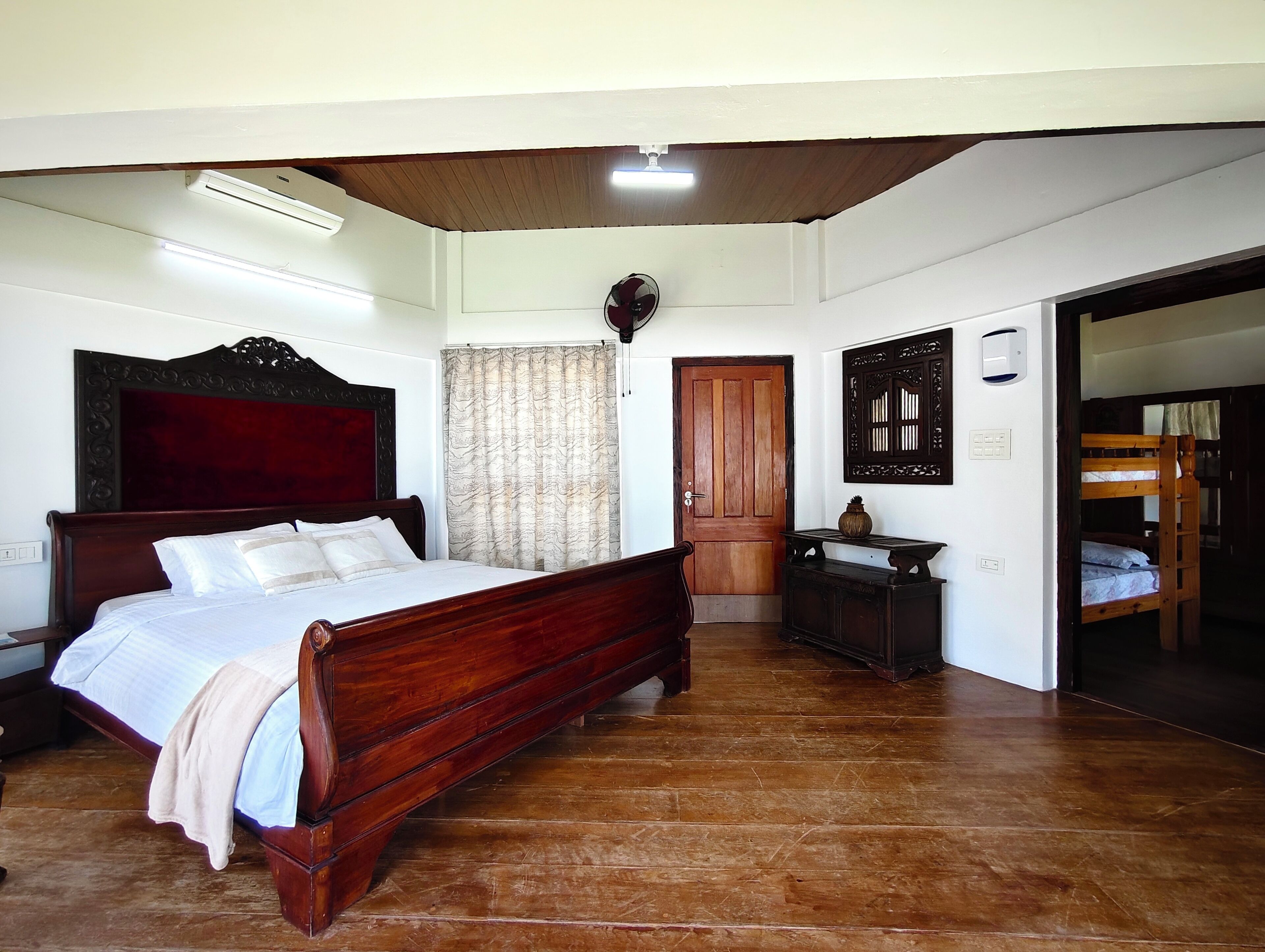 Quarto com uma cama de madeira, um criado-mudo, uma cômoda, uma porta e vista para uma cama de solteiro no quarto vizinho.