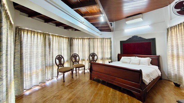 A bedroom with a wooden bed, two chairs, a large window with curtains, and a wooden ceiling. - Cheshire Village - Beach Resort (Kannur)
