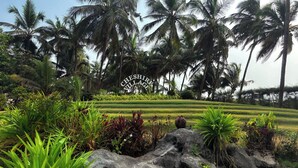 A lush garden with palm trees and a sign reading "Cheshire Village". - Cheshire Village - Beach Resort (Kannur)