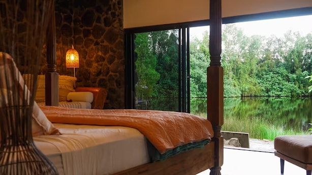 Une chambre avec une grande fenêtre, un lit recouvert d’une couverture orange, un mur en pierre et un luminaire suspendu.