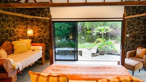 Une chambre avec un grand lit, une tête de lit en bois, un mur en pierre et une vue sur un étang et la verdure environnante.
