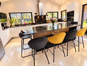 A modern kitchen with a central island, bar stools, and a view of the outdoors. - Villa Les Chênes Verts – Private Pool – Hot Tub – Provence – Sleeps 10 (Sainte-Anastasie-sur-Issole)