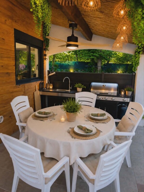Une table de salle Ă manger ronde, entourĂ©e de chaises blanches, avec un barbecue et une vue sur la verdure par la fenĂȘtre.