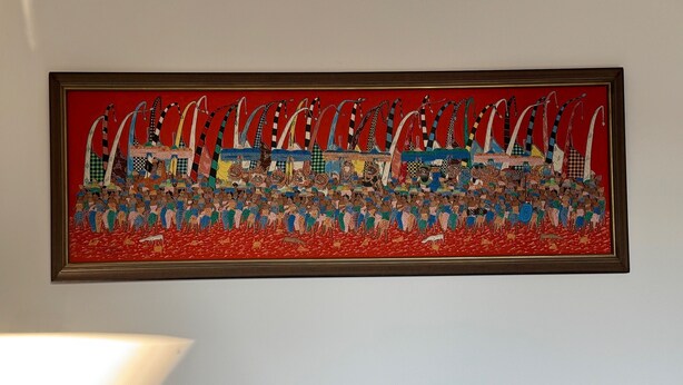 Une œuvre d’art encadrée représentant une scène animée avec de nombreuses figures et des bateaux sur un fond rouge.